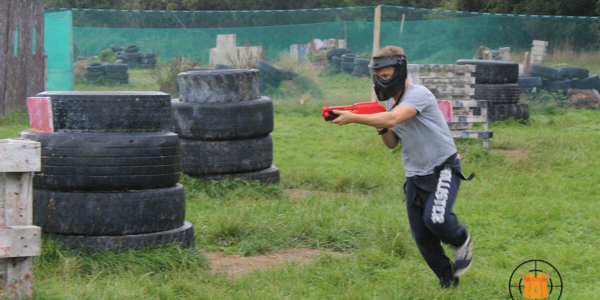 Stag Party Kilkenny A boy shooting a splatball gun and wearing a safety mask runs through a paintball arena with tyre walls and timber obstacles.
