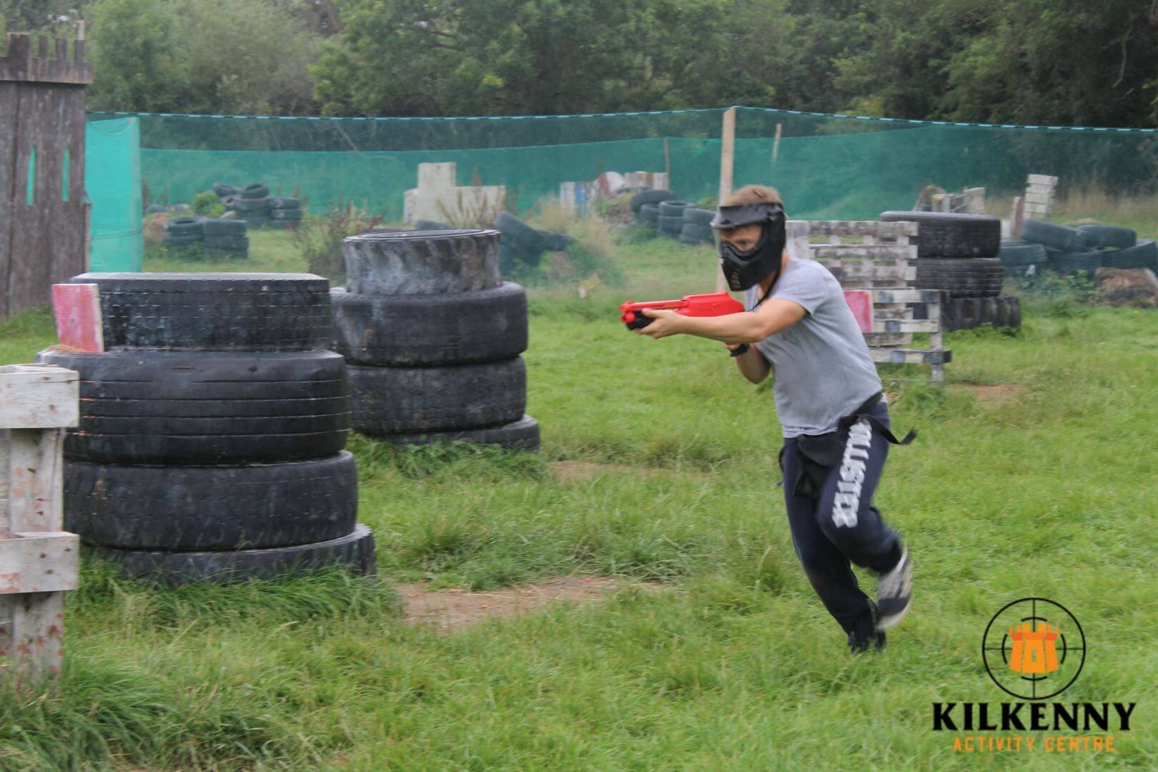 Stag Party Kilkenny A boy shooting a splatball gun and wearing a safety mask runs through a paintball arena with tyre walls and timber obstacles.